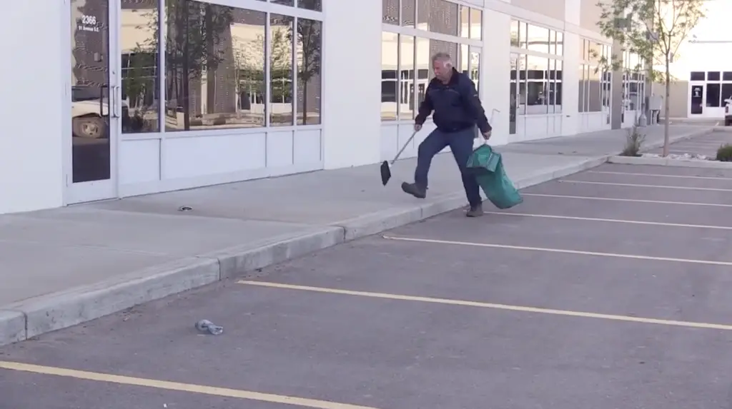  Brian is walking through a parking lot collecting litter with a tool and a rubbish bag in hand.